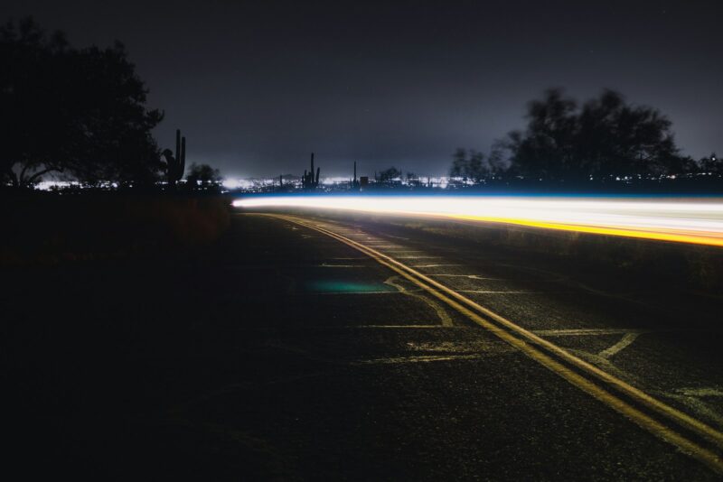 Long exposure of a car's headlights and tail lights creating streaks of light along a dark, empty road at night, with city lights in the far distance.