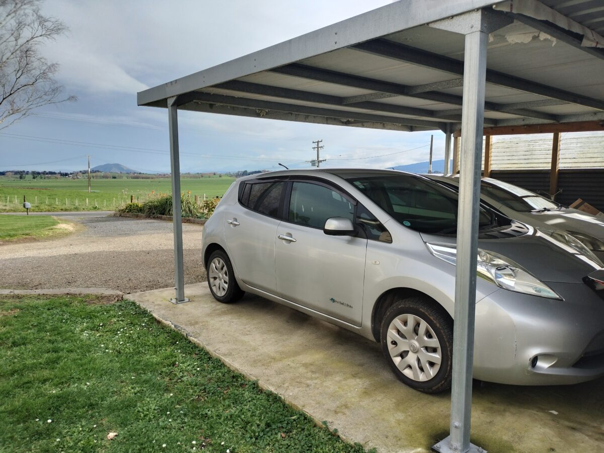 Silver car parked under a metal carport, with a rural landscape backdrop. Silver car parked under a metal carport, with a rural landscape backdrop.