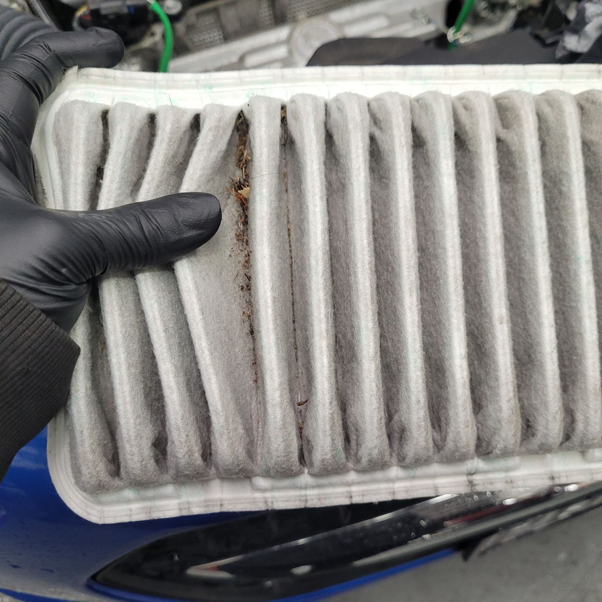 Used Engine Air Filter with Debris Hand wearing black glove holding a dirty pleated air filter with debris trapped inside.
