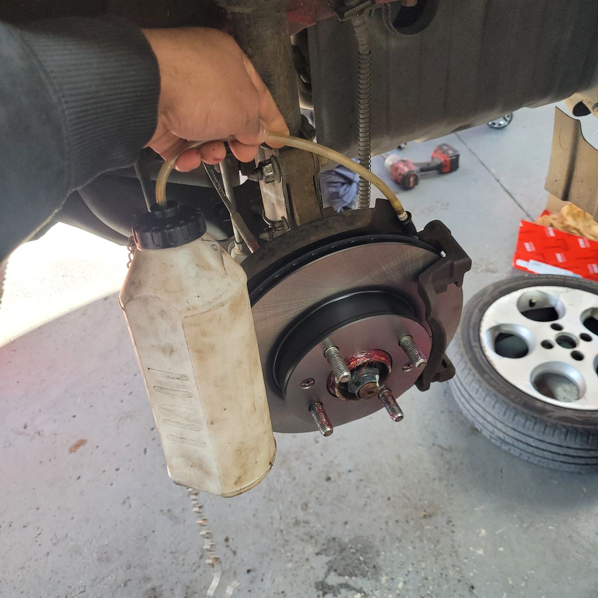 Hand performing brake fluid flush with bottle and hose attached to car brake caliper.