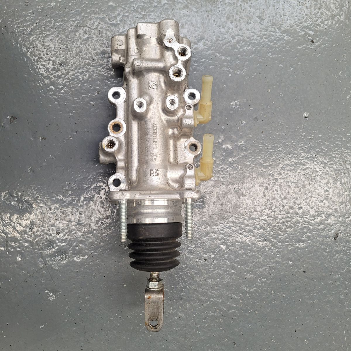 Automotive Brake Master Cylinder on Workshop Floor A metallic brake master cylinder with mounts and plastic nozzles on a gray floor.