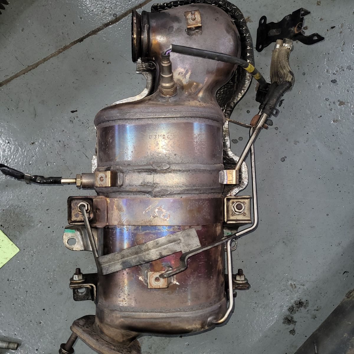 Used Automotive Catalytic Converter on Workshop Floor A metal catalytic converter with sensors and pipes, lying on a gray workshop floor.