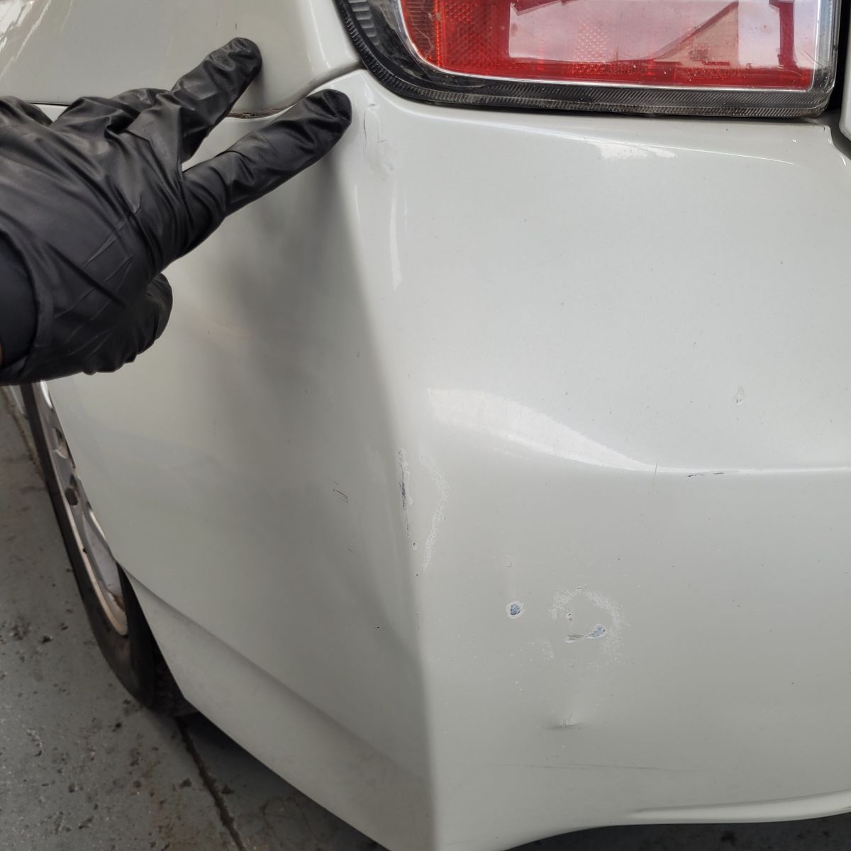 Inspecting Rear Bumper Damage on White Car Gloved hand pointing at dent and scratches on white car bumper near taillight.