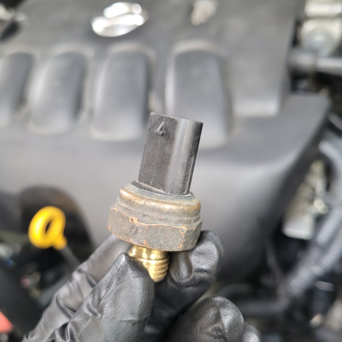 A gloved hand holding a coolant temperature sensor in front of a car engine.