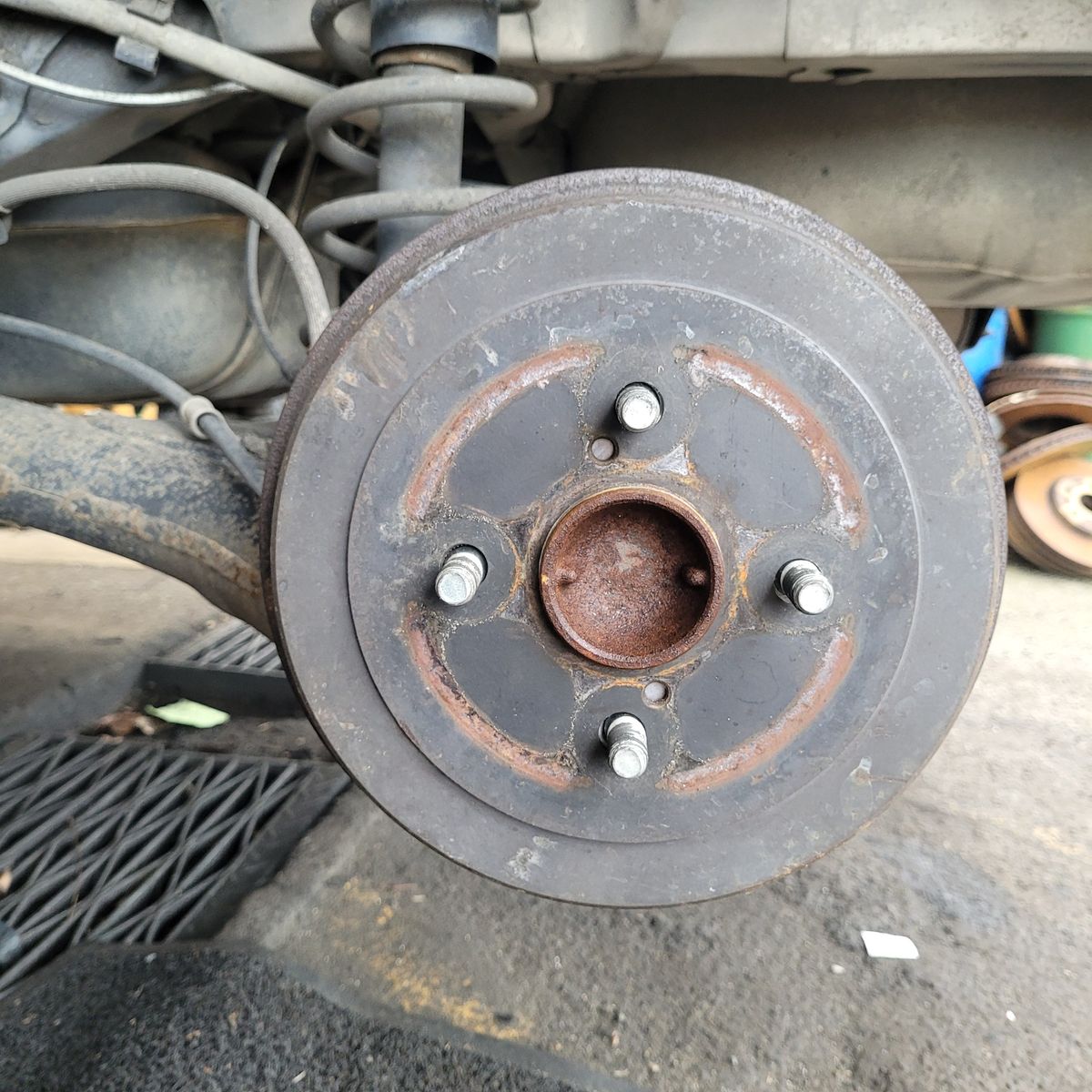 Car drum brake showing five wheel studs and rusty surface, attached to suspension.