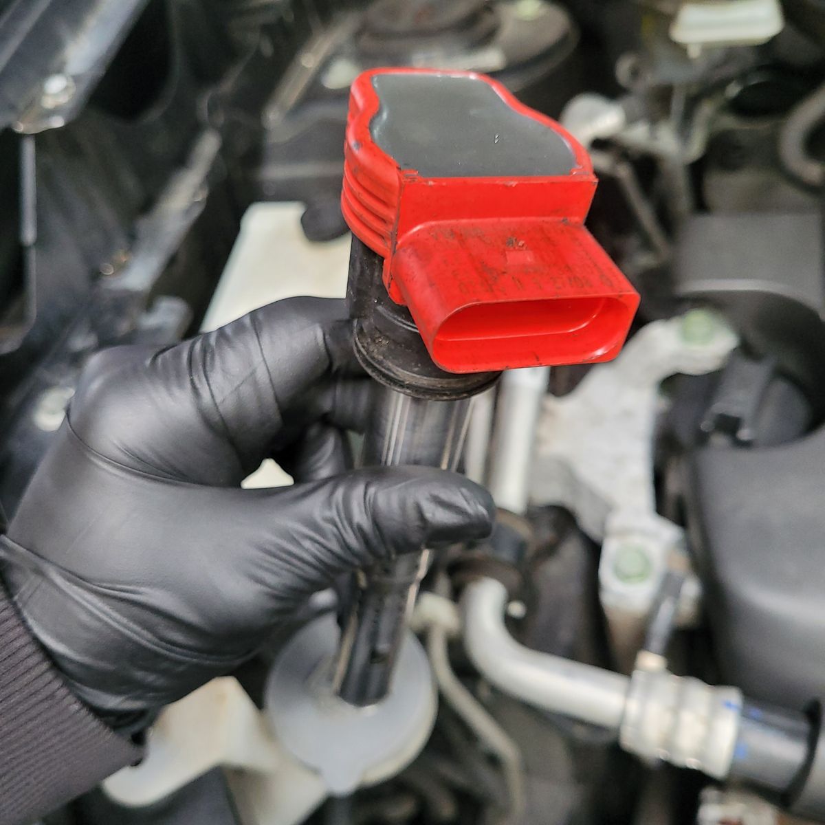 Gloved hand holding a red electronic ignition coil above an engine bay