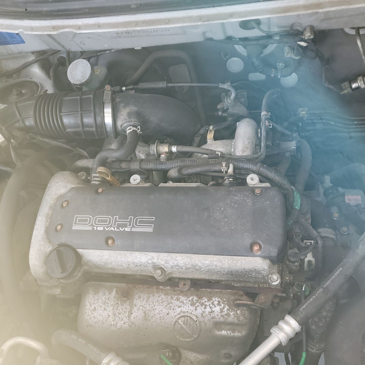 DOHC 16 Valve Car Engine Close-Up Overhead view of a car engine labeled DOHC 16 Valve with visible hoses and components.