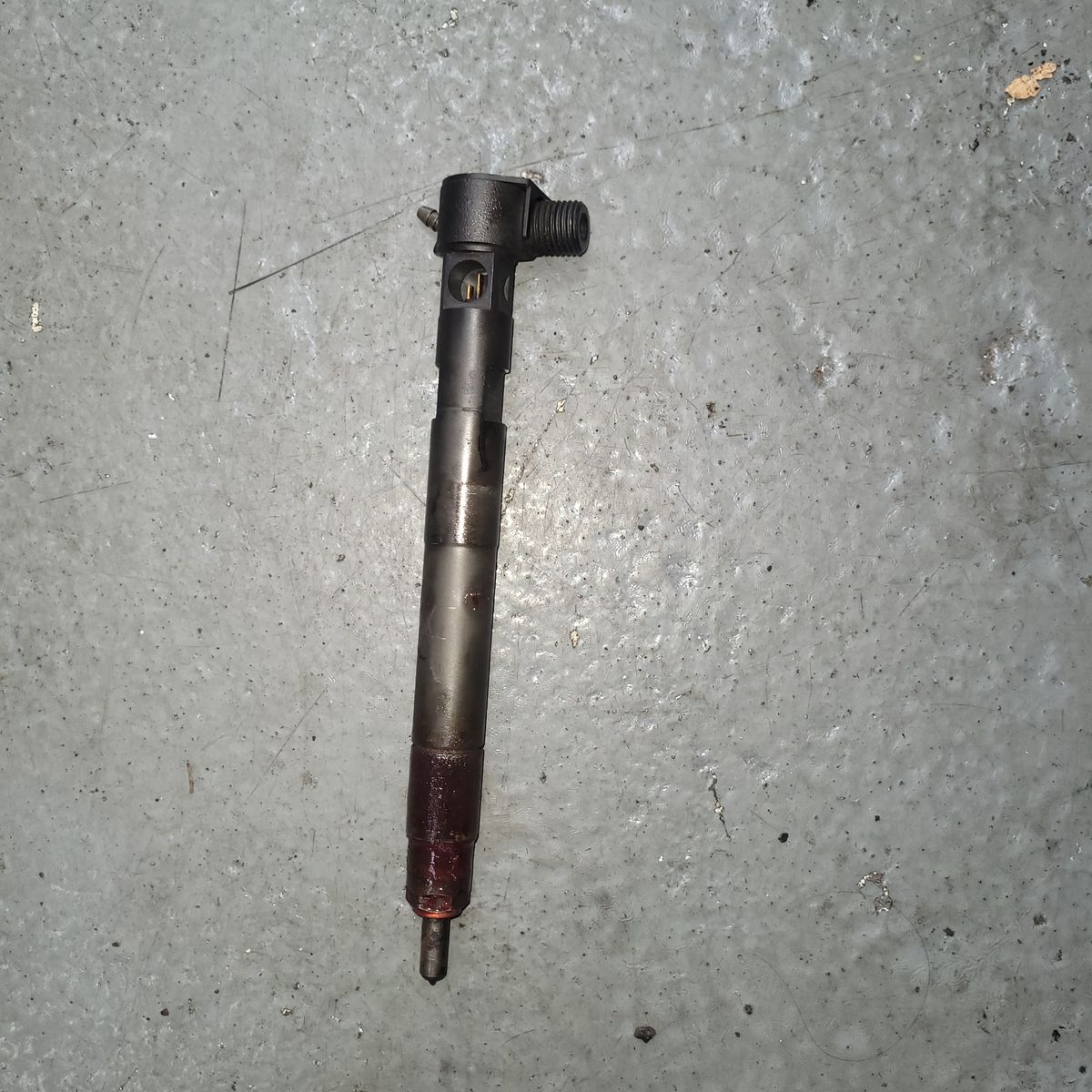 Used Fuel Injector on Workshop Floor A single fuel injector lying on a grey, textured workshop floor.