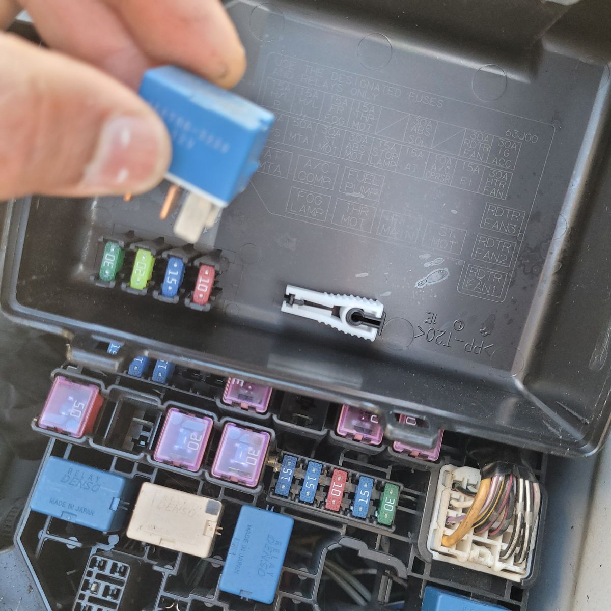 Person holding blue fuel pump relay near vehicle’s fuse box and diagram.