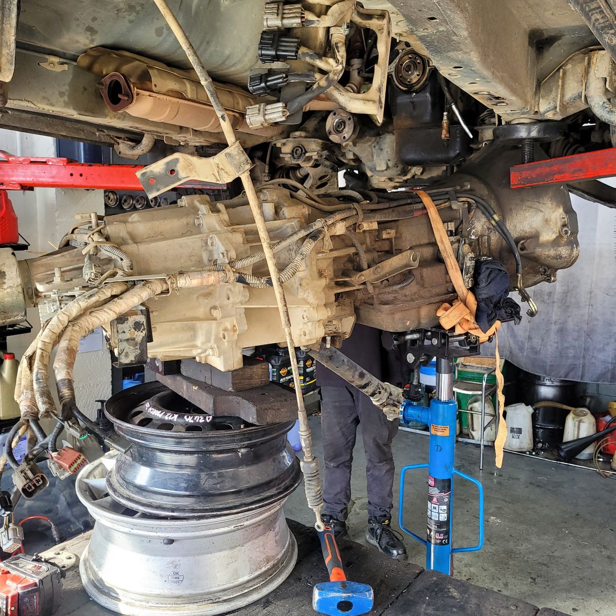 Disassembled Vehicle Gearbox on Lift in Workshop A car gearbox is being removed underneath a lifted vehicle using a jack and old wheel rims for support.