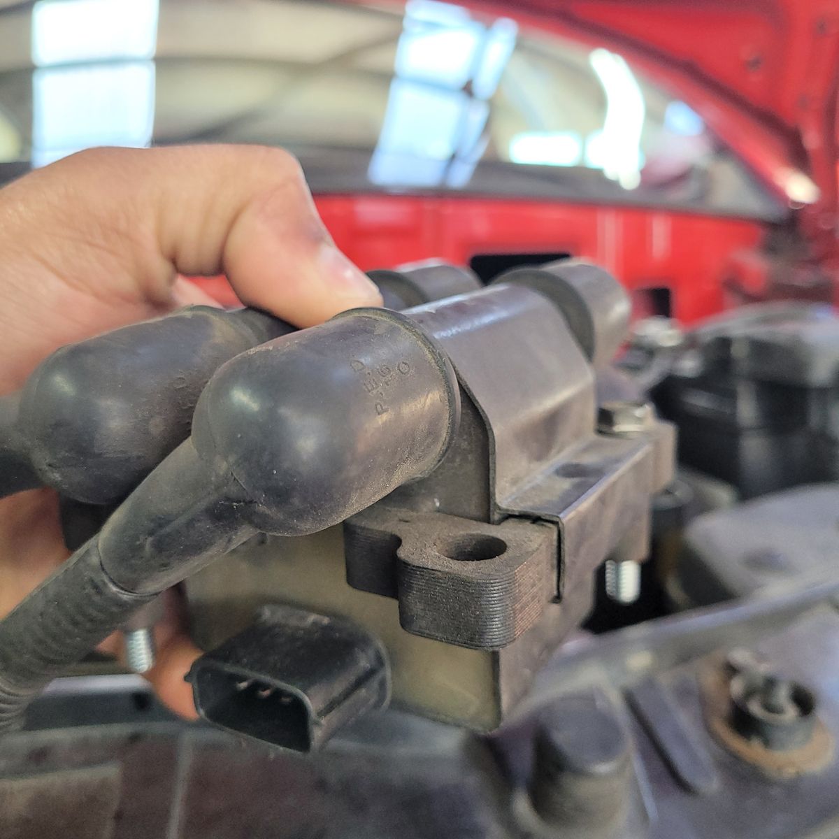 Car Ignition Coil Close-Up Hand holding a car ignition coil with attached spark plug wires inside an engine bay.