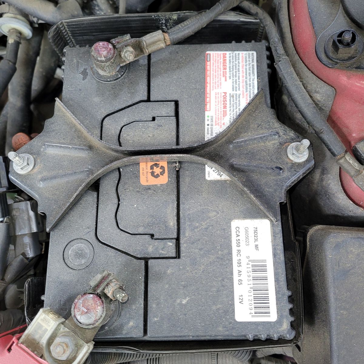 Dusty 12V car battery secured by a metal bracket in an engine compartment.