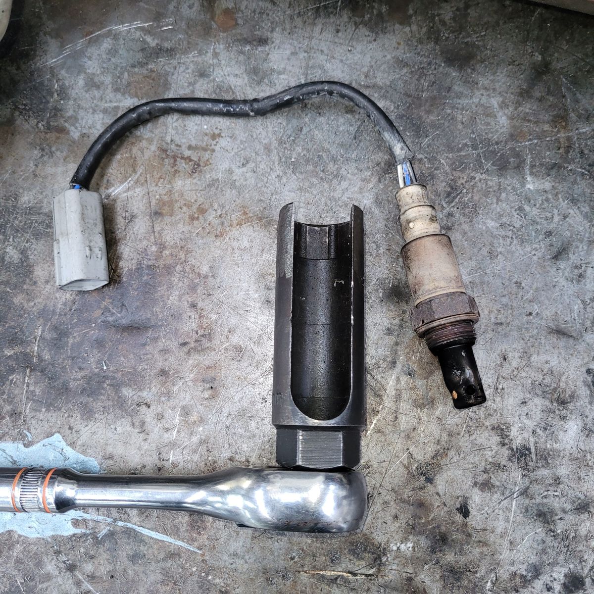 Oxygen Sensor and Socket Wrench on Workbench A used oxygen sensor and socket wrench placed on a dirty metal surface.