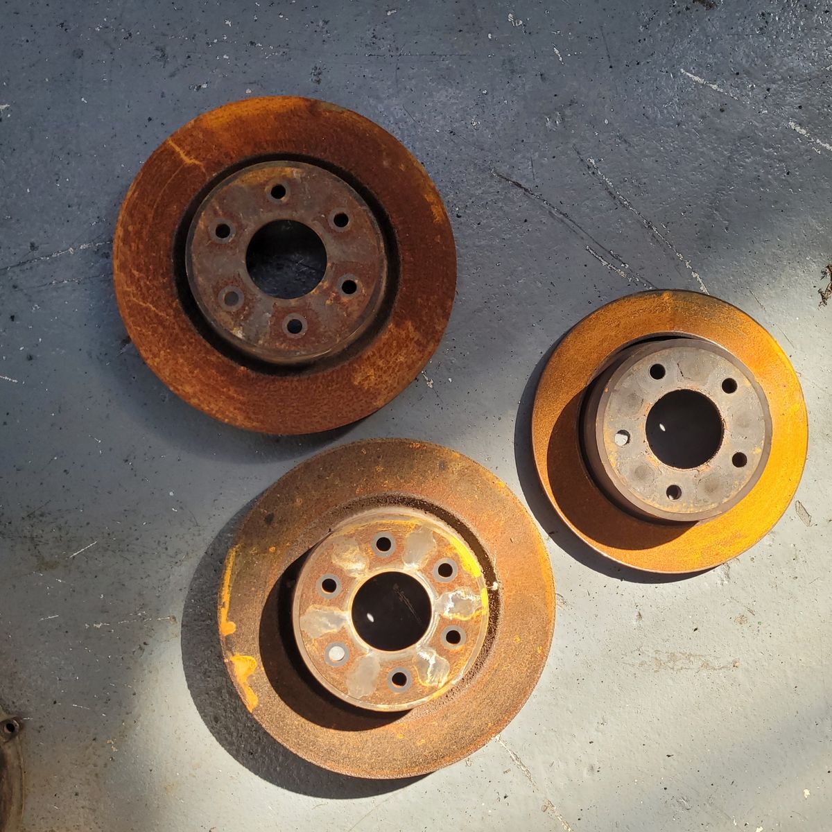 Three Rusty Brake Discs on Workshop Floor Three rusty brake discs lying on a grey workshop floor in sunlight.
