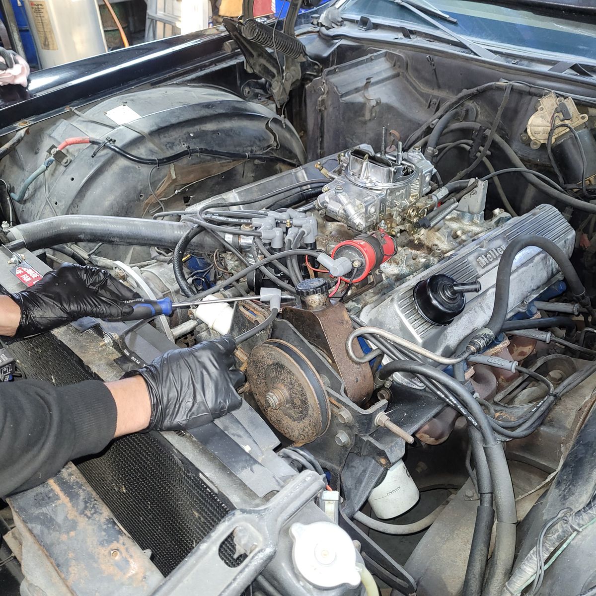 Gloved hands using tools to work on a car engine in an open hood.